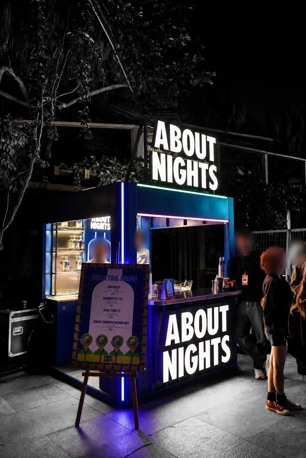 Gece etkinliğinde ışıklı About Nights barı; parlayan harfler, mavi LED çizgiler ve servis yapan bar ekibi.