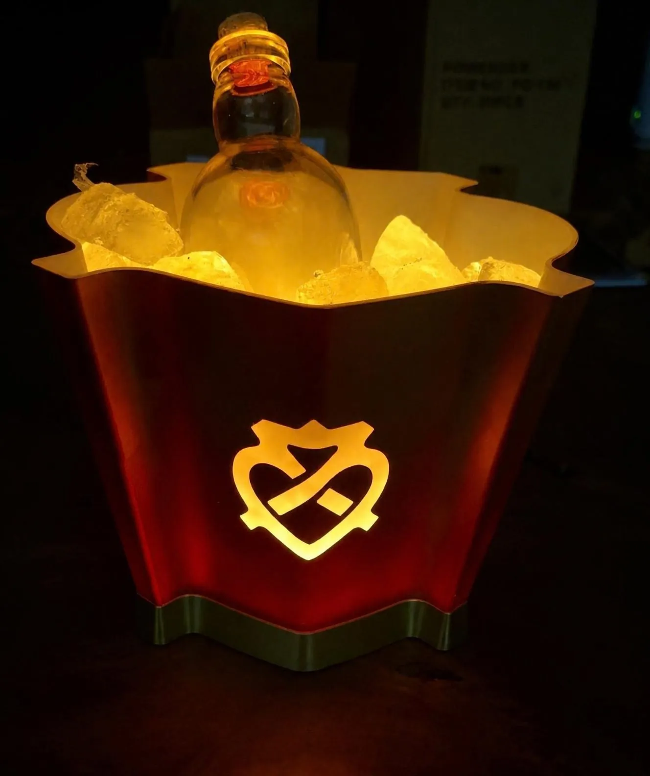 Gespritzter Eiskübel, bernsteinfarben leuchtend, mit beleuchtetem Logo und einer Flasche im Eis vor schwarzem Hintergrund.