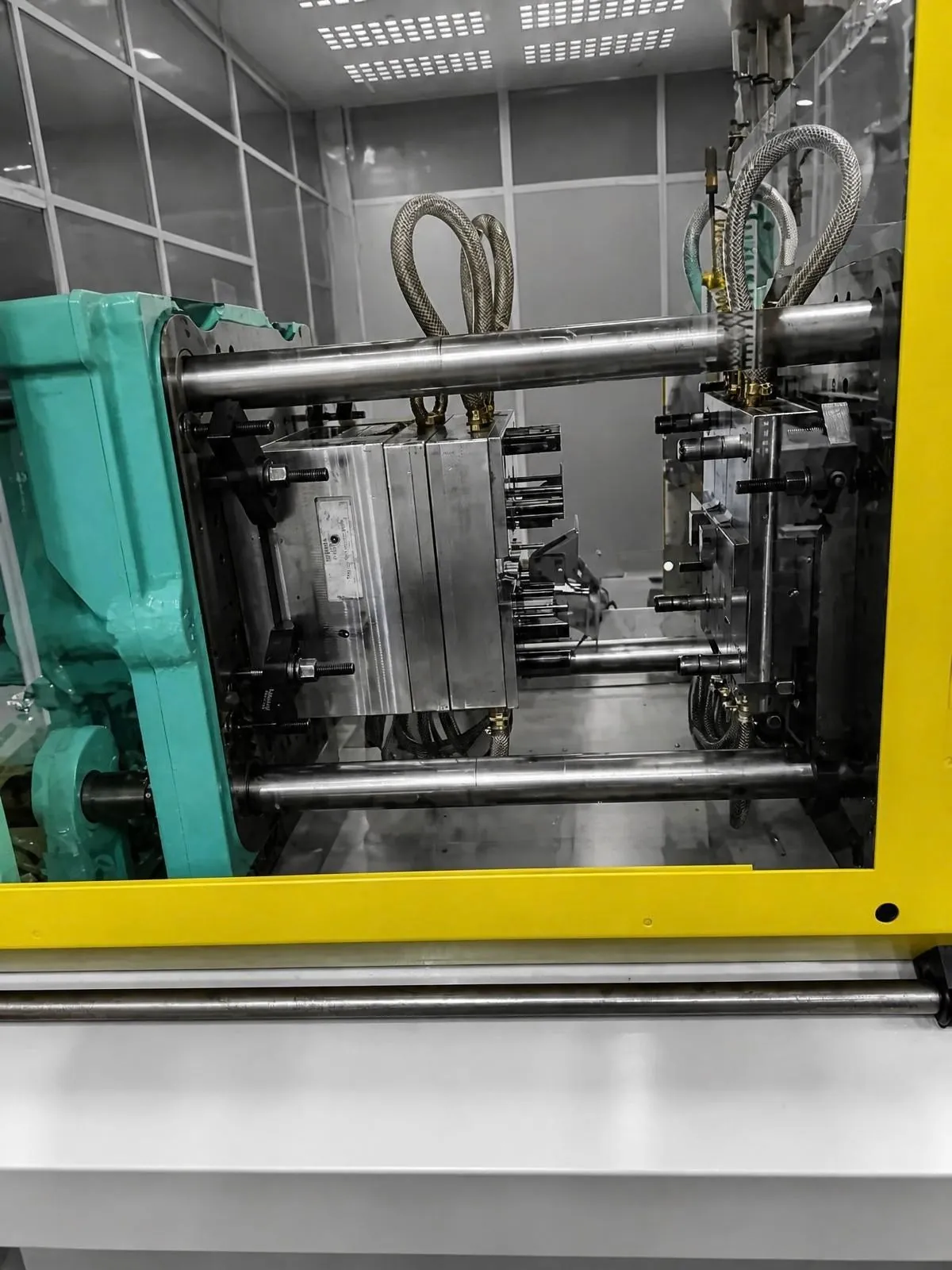 Spritzgießmaschine mit gelbem Schutzrahmen, Schließeinheit und Hydraulikleitungen, von der Bedienseite aus gesehen.