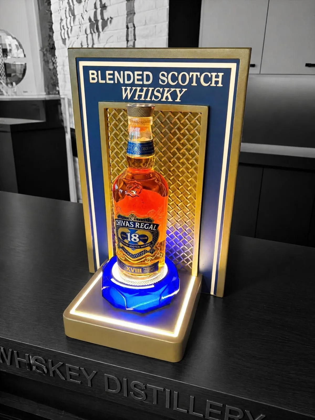 Counter display with a gold perforated back panel, a lit base and a brand header holding one bottle.