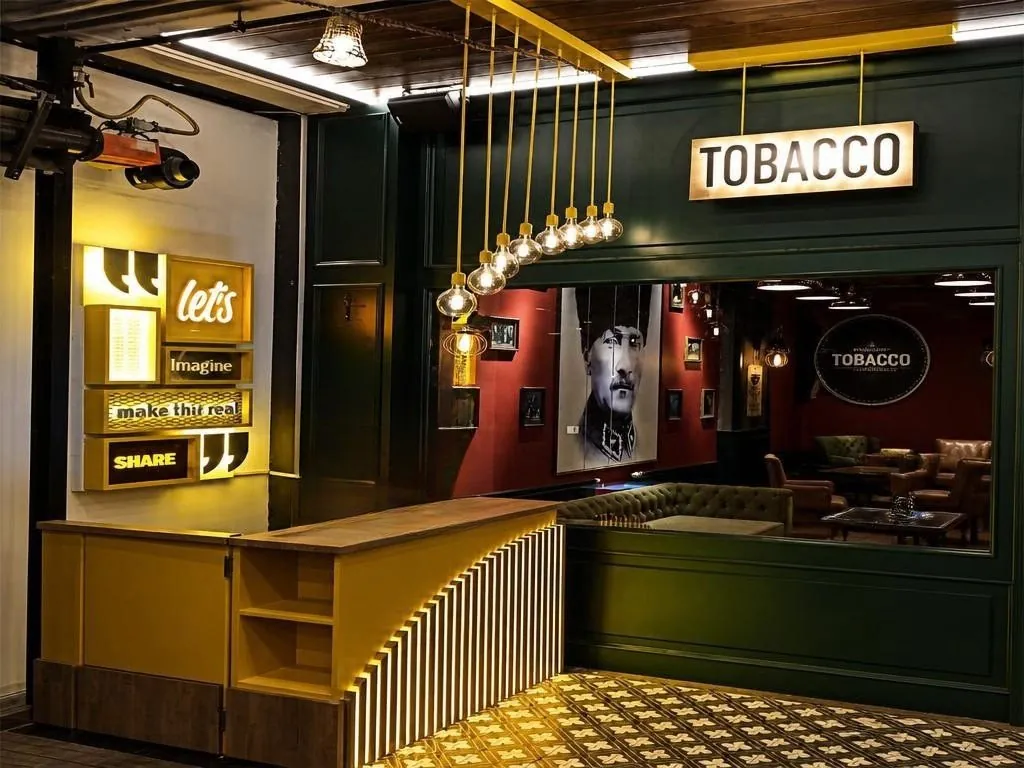Ladeninnenraum in Dunkelgrün und Messing mit „Tobacco"-Leuchtschild, Pendelleuchten und gemustertem Boden.
