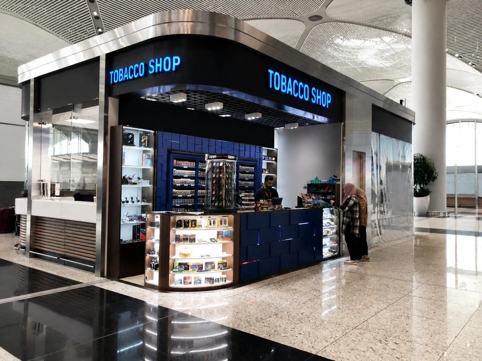 Blaue „Tobacco Shop"-Einheit in einem Flughafenterminal mit beleuchteten Warenwänden und Edelstahlvordach.