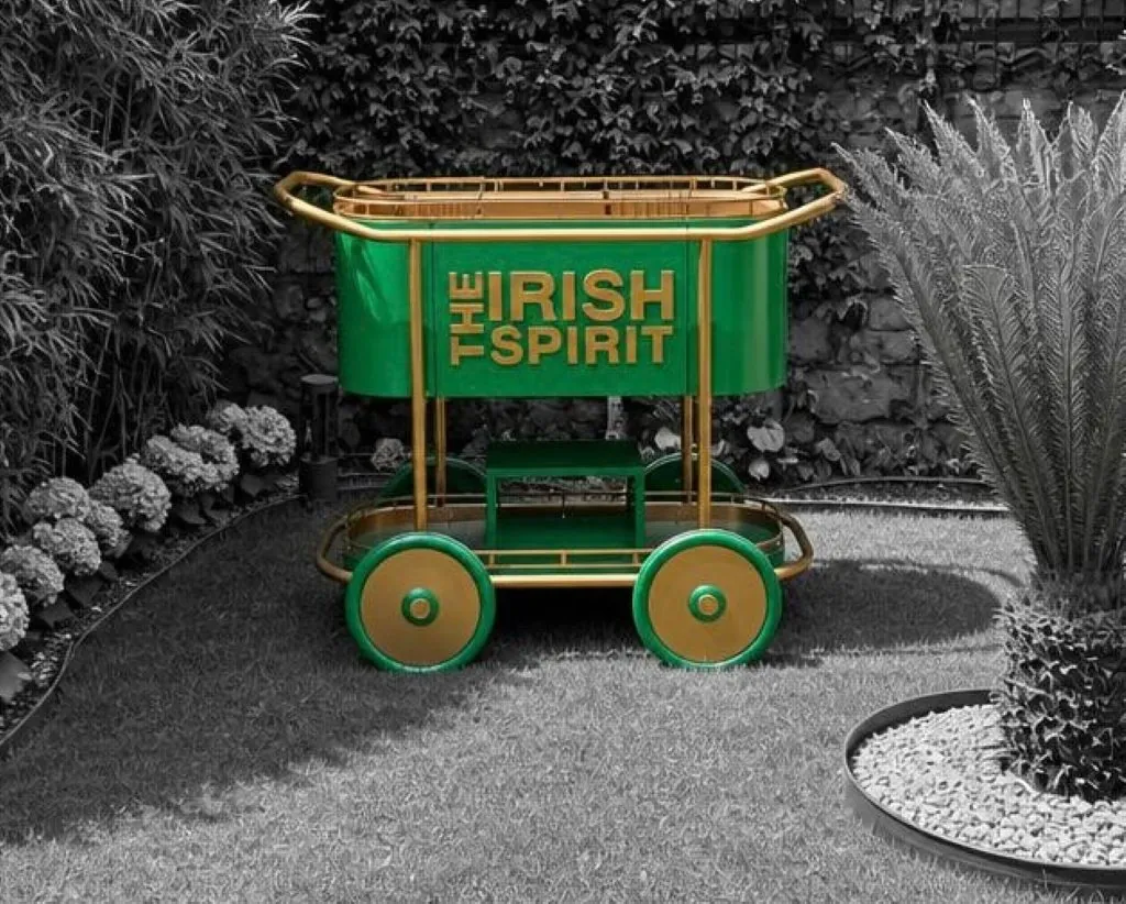 Grün-goldener Servierwagen auf Speichenrädern mit Markenschriftzug, aufgestellt in einem Garten.