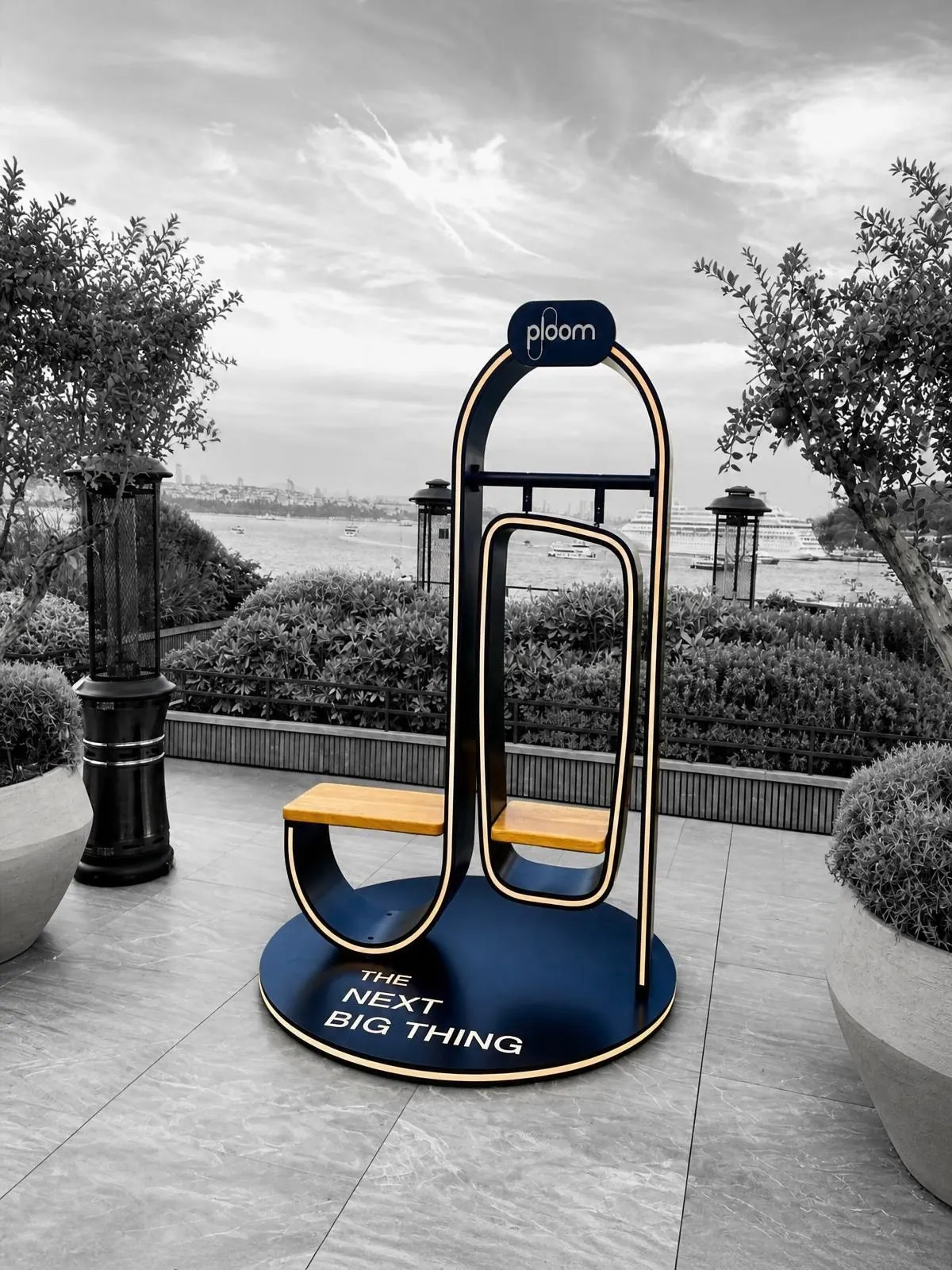 Sculptural structure with a navy arch and a wooden seat, standing on a round navy base with a brand plate, on a rooftop terrace overlooking the Bosphorus.